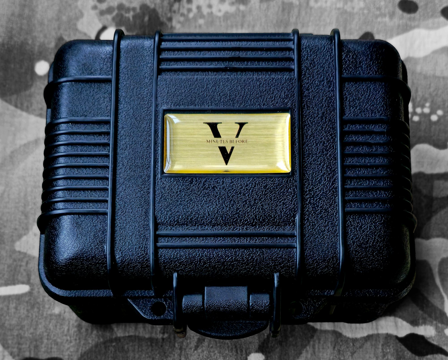 A black, rugged case with a ribbed texture and a gold V plate, resembling the Five Minutes Befores NORMANDY Field Watch 36mm, rests closed on a camouflage-patterned surface, echoing its field watch durability.