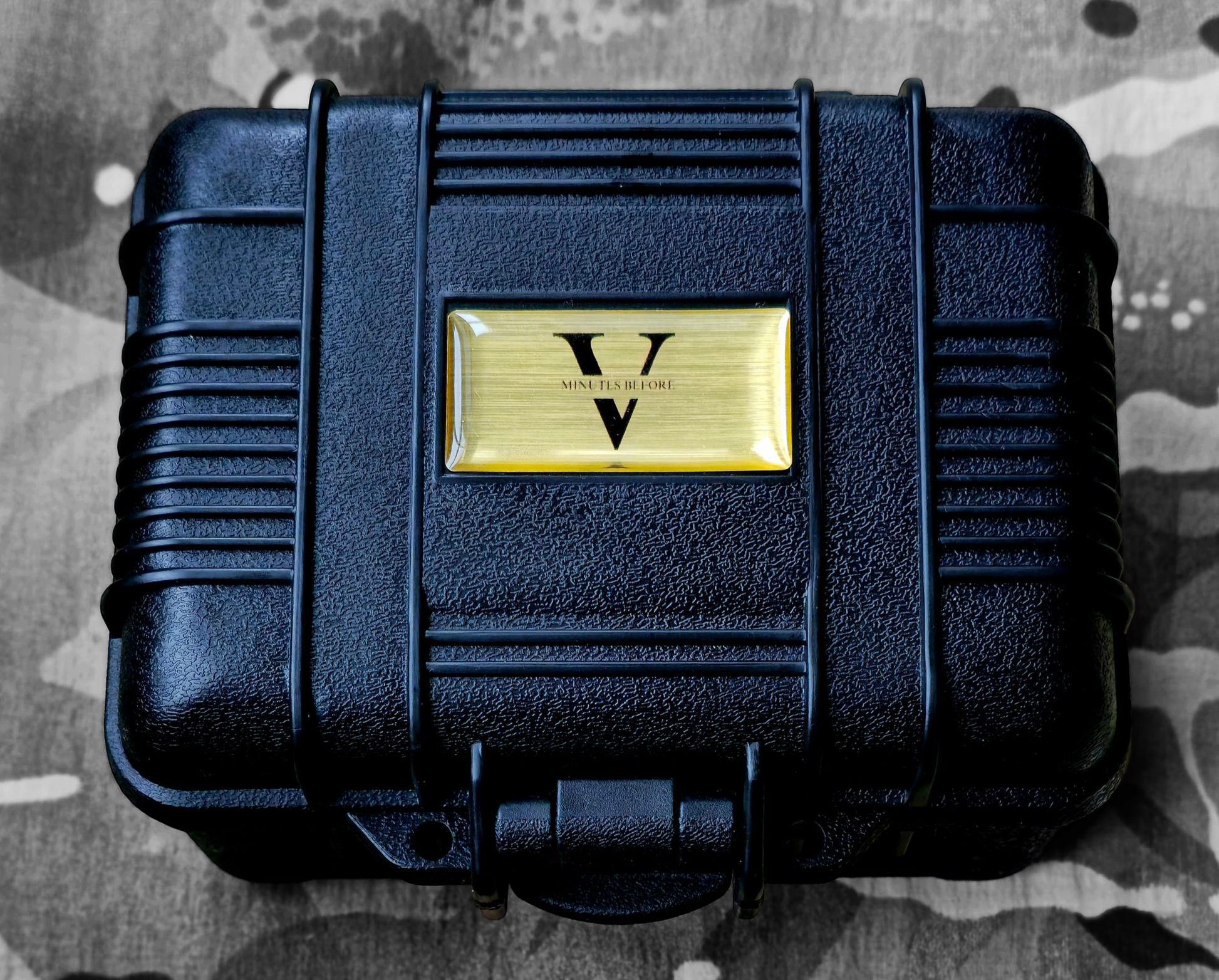 A black, rugged case with a ribbed texture and a gold V plate, resembling the Five Minutes Befores NORMANDY Field Watch 36mm, rests closed on a camouflage-patterned surface, echoing its field watch durability.