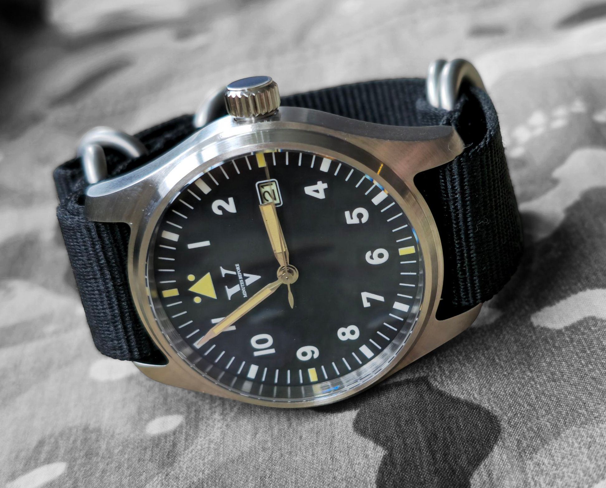 The VANGUARD Field Watch by Five Minutes Before features a 39mm stainless steel case, black dial with white Arabic numbers, a luminous glow, date window, and a black fabric strap on a camouflage surface.