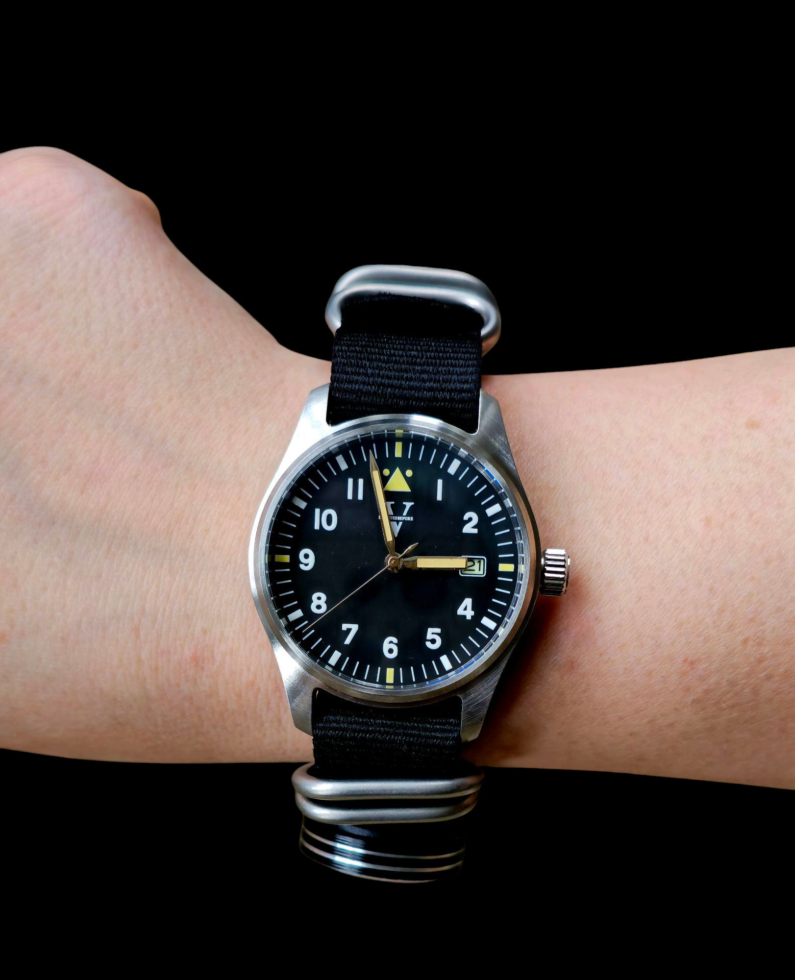 The wrist sports a Five Minutes Before VANGUARD (Field Watch 39mm) featuring a stainless steel case, black dial, white numerals, and yellow second hand. Its black fabric strap with silver hardware adds a luminous touch against the dark backdrop.