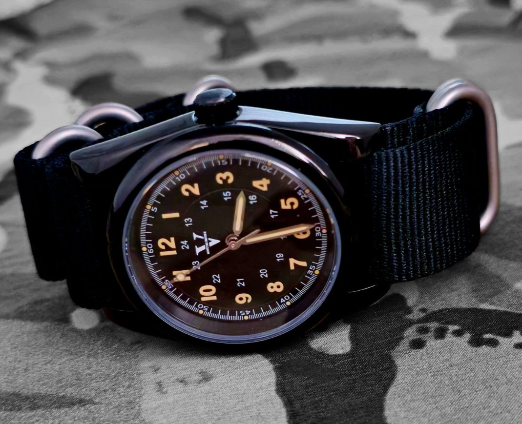 The Five Minutes Before ECLIPSE Field Watch 36mm boasts a polished stainless steel case and a dark dial with white numbers and hands. Its sleek design on a black nylon strap, set against a blurred camo background, creates an elegant silhouette akin to an eclipse.