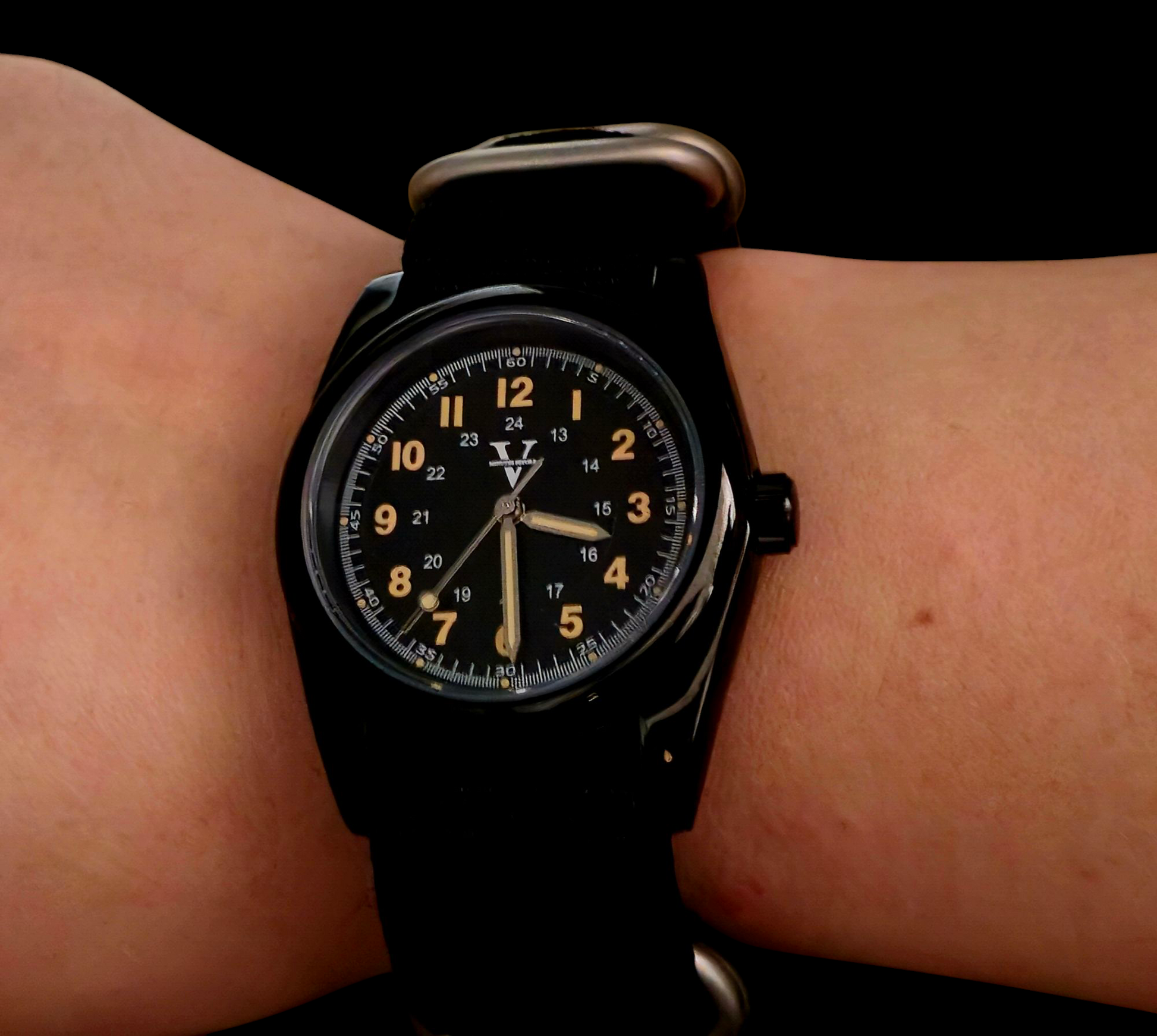 A person wearing the ECLIPSE (Field Watch 36mm) by Five Minutes Before showcases both 12-hour and 24-hour formats with yellow numerals on a round face. The watch has a black fabric band, a single crown, and a stainless steel case, all set against a dark background reminiscent of an eclipse.